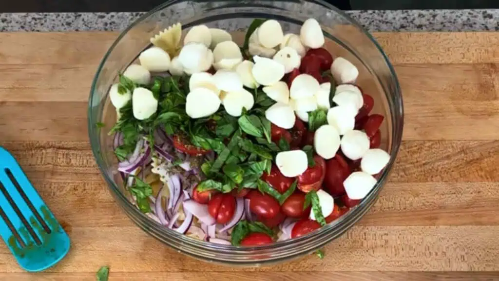 A glass bowl filled with sliced red onion, grape tomatoes, mozzarella balls, fresh basil, and pasta on a wooden counter.
