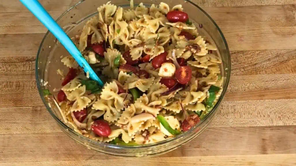 A glass bowl of bowtie pasta salad with cherry tomatoes, green vegetables, and a blue serving spoon on a wooden surface.