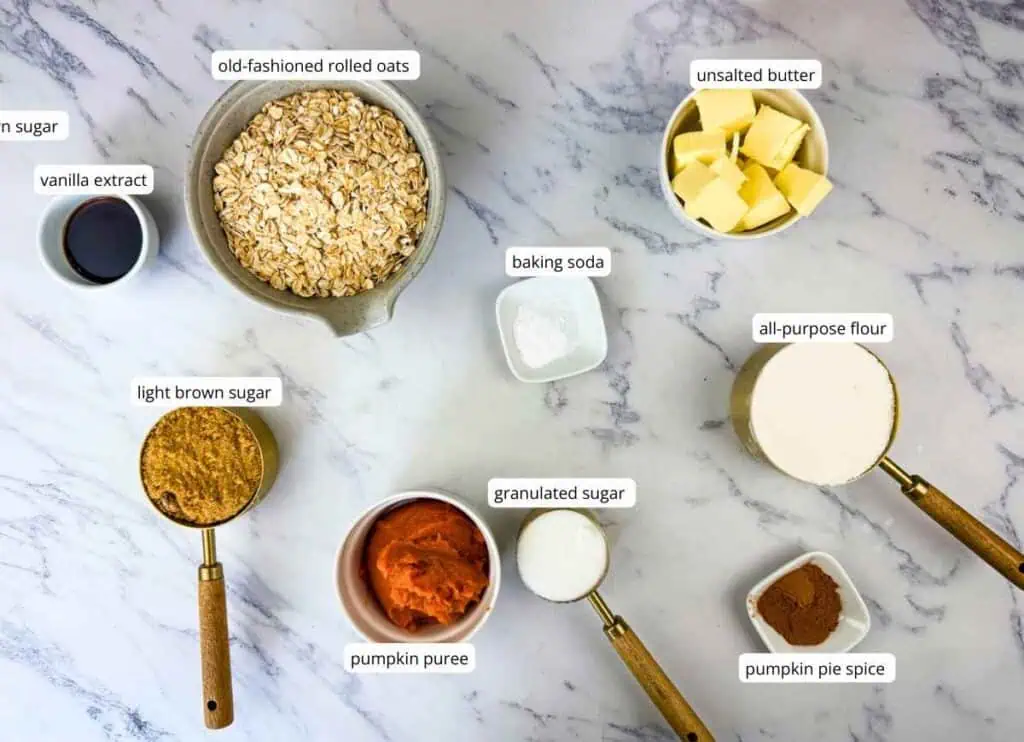 Baking ingredients on a marble surface include oats, butter, light brown sugar, pumpkin puree, granulated sugar, flour, pie spice, baking soda, and vanilla extract, each labeled.