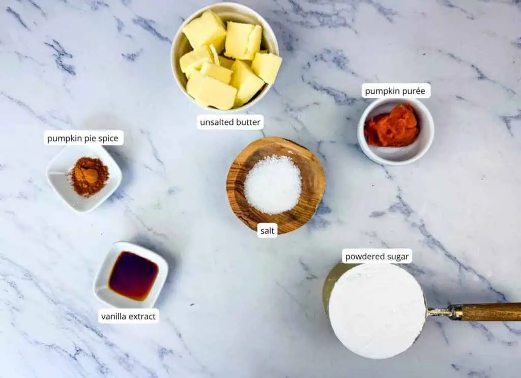 Six ingredients on a marble surface: unsalted butter, pumpkin purée, pumpkin pie spice, vanilla extract, salt, and powdered sugar, each in a separate container with labels.