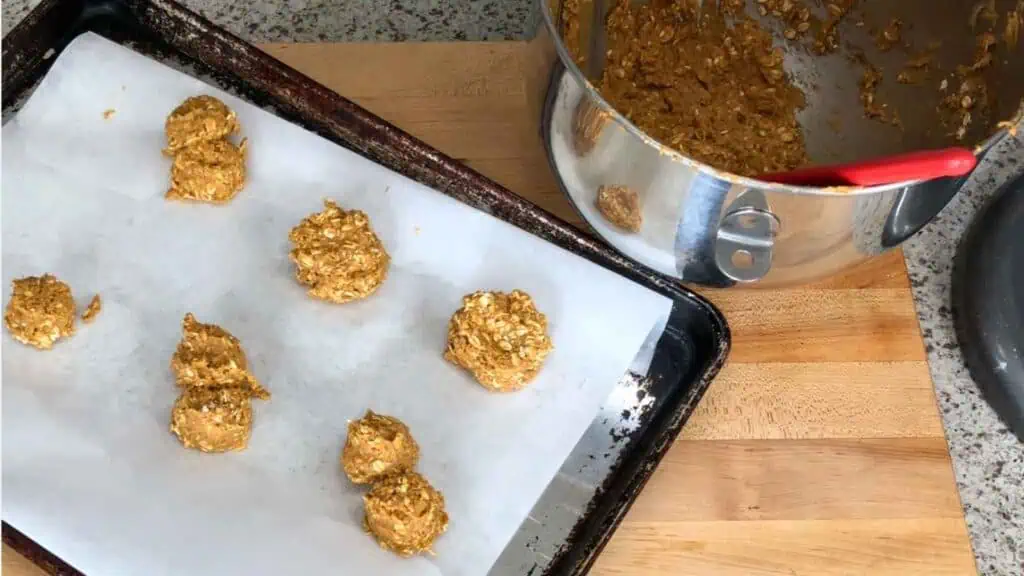 Unbaked cookie dough scoops on a parchment-lined baking sheet with a mixing bowl and spatula nearby on a kitchen counter.