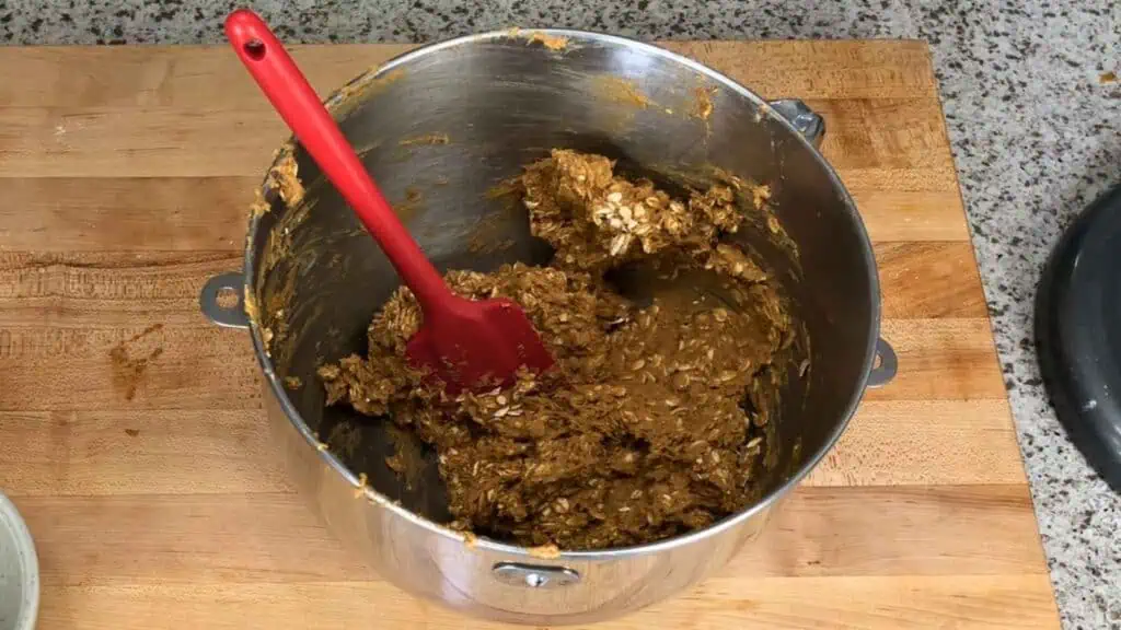 A red spatula rests in a metal mixing bowl containing an oat and brown batter, sitting on a wooden surface next to a granite countertop.