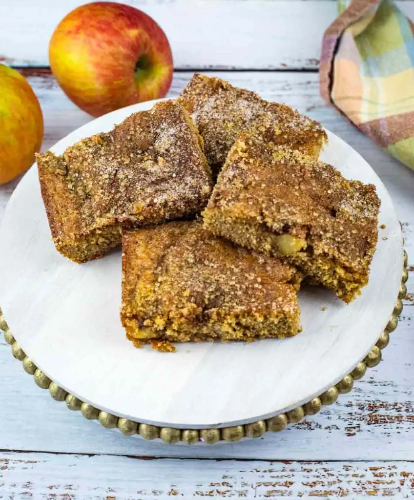 Four slices of apple cinnamon cake are arranged on a white cake stand, with whole apples and a cloth napkin in the background.