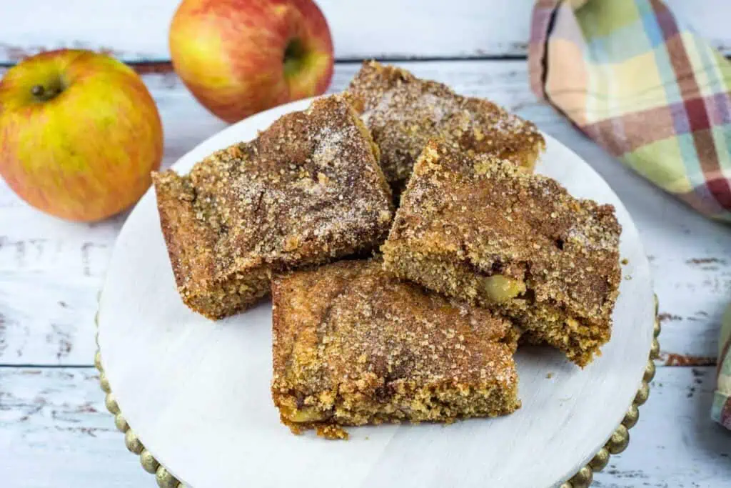 Four brown apple cake squares are arranged on a white plate with two whole apples and a plaid cloth in the background, all set on a rustic wooden surface.