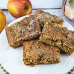 Four brown apple cake squares are arranged on a white plate with two whole apples and a plaid cloth in the background, all set on a rustic wooden surface.