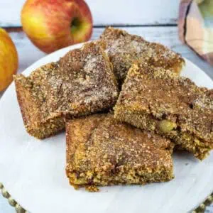 Four brown apple cake squares are arranged on a white plate with two whole apples and a plaid cloth in the background, all set on a rustic wooden surface.