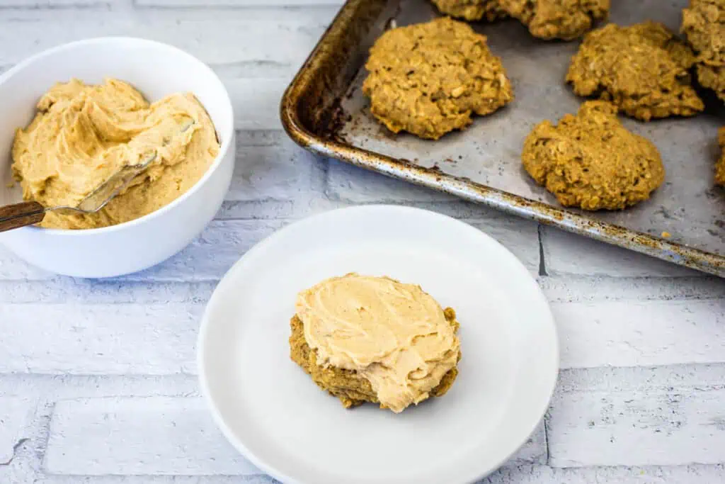 A plate with a frosted cookie, a bowl of frosting with a spoon, and a baking sheet with unfrosted cookies on a white surface.