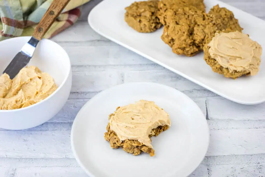 A partially eaten cookie with frosting sits on a plate, with more cookies on a tray and a bowl of frosting and a spreading knife nearby.
