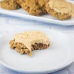 A frosted cookie with a bite taken out sits on a white plate, with more cookies on a platter in the background.