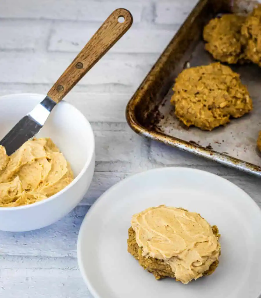 A frosted cookie sits on a white plate next to a bowl of frosting with a spatula and a baking sheet with more cookies on a white surface.