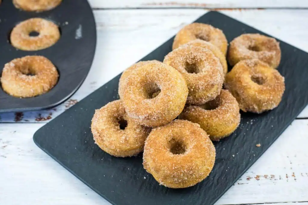 A black platter holds a stack of small cinnamon sugar donuts. A donut baking tray with more donuts is partially visible on the left.