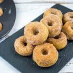 A black platter holds a stack of small cinnamon sugar donuts. A donut baking tray with more donuts is partially visible on the left.