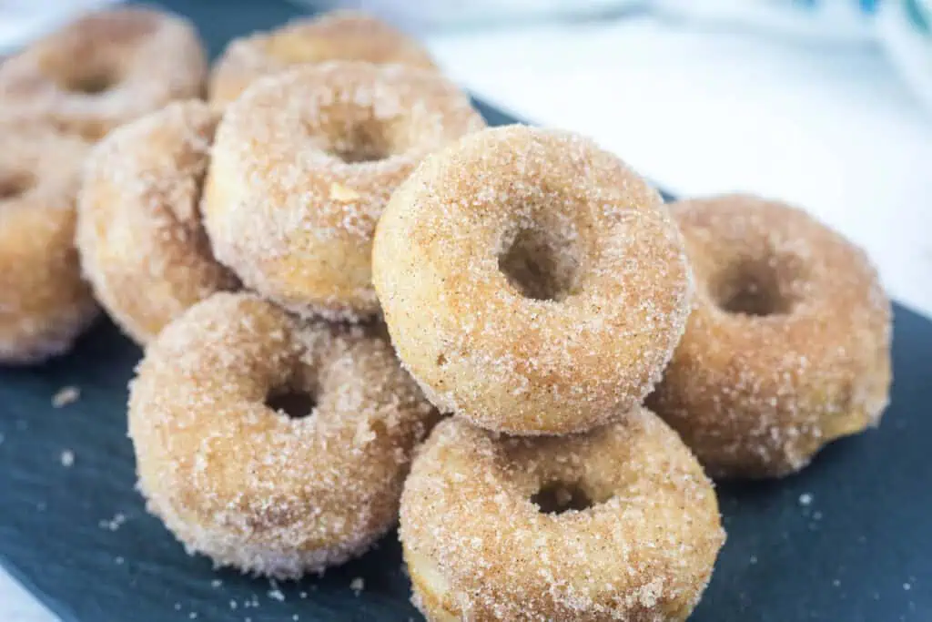 A pile of sugar-coated baked donuts is arranged on a dark serving tray, with a light background visible behind them.