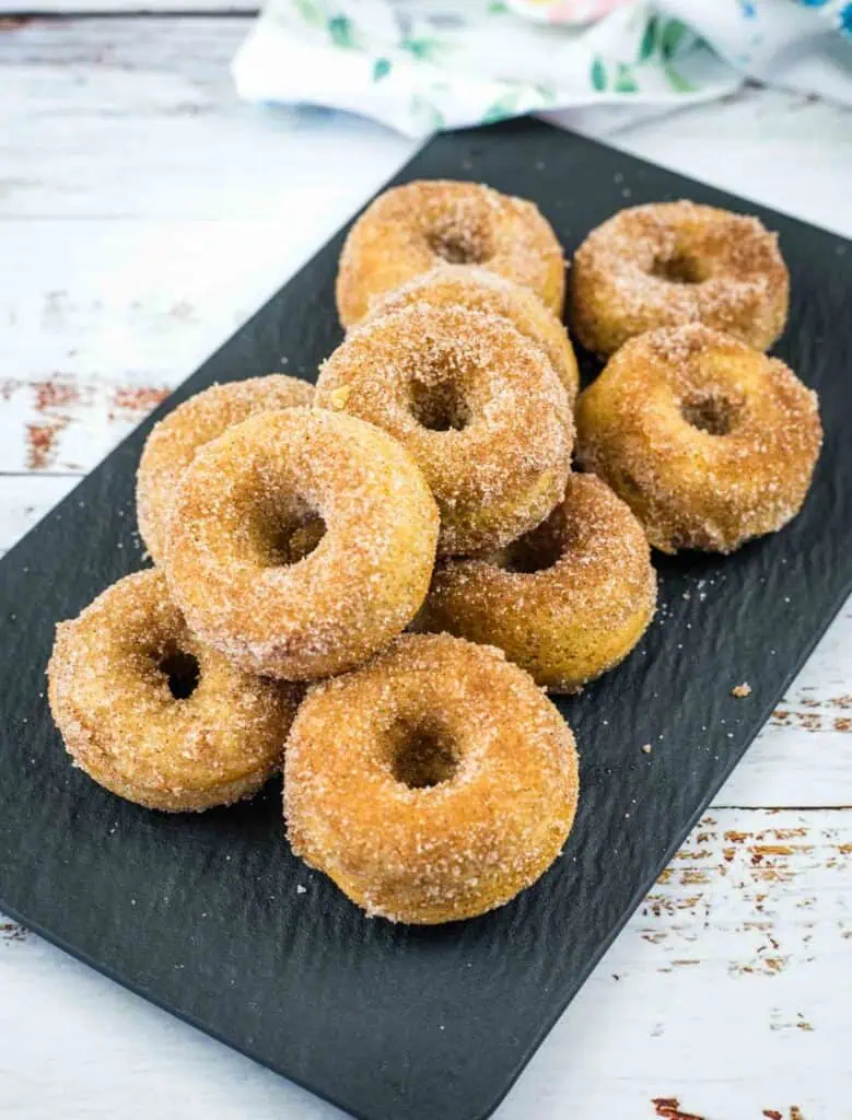 A black rectangular plate holds a pile of cinnamon sugar-coated donuts on a rustic white wooden surface.