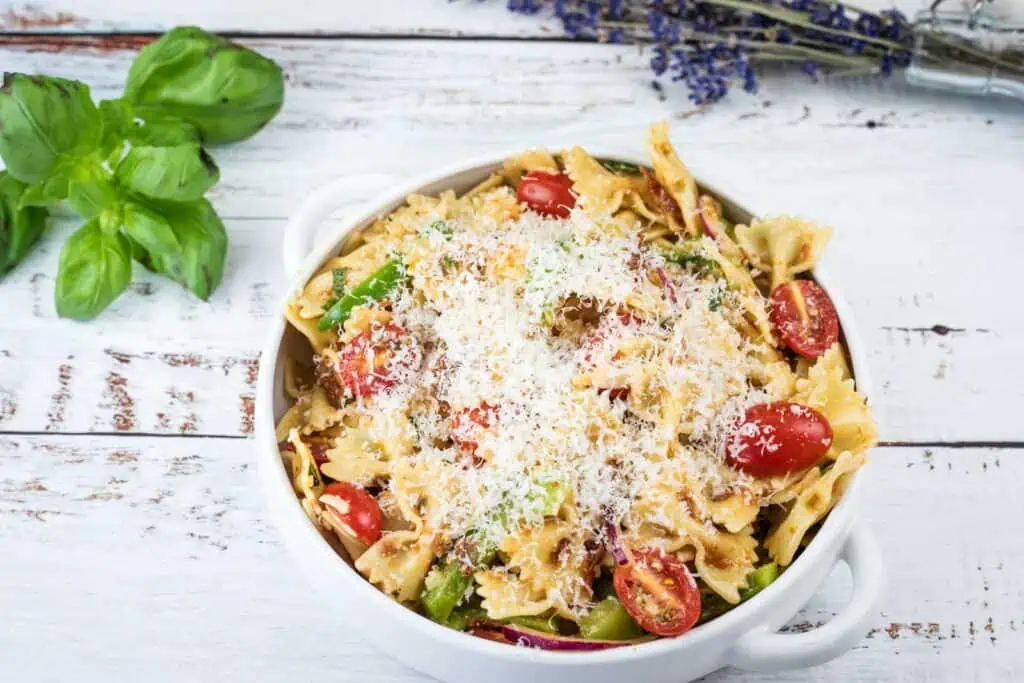 Bowl of farfalle pasta salad with cherry tomatoes, asparagus, and grated cheese on top, set on a white wooden table with fresh basil nearby.
