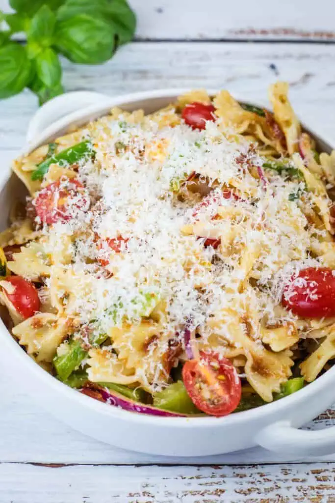 Bowl of farfalle pasta salad with cherry tomatoes, green vegetables, red onion, and topped with grated cheese on a white wooden surface.