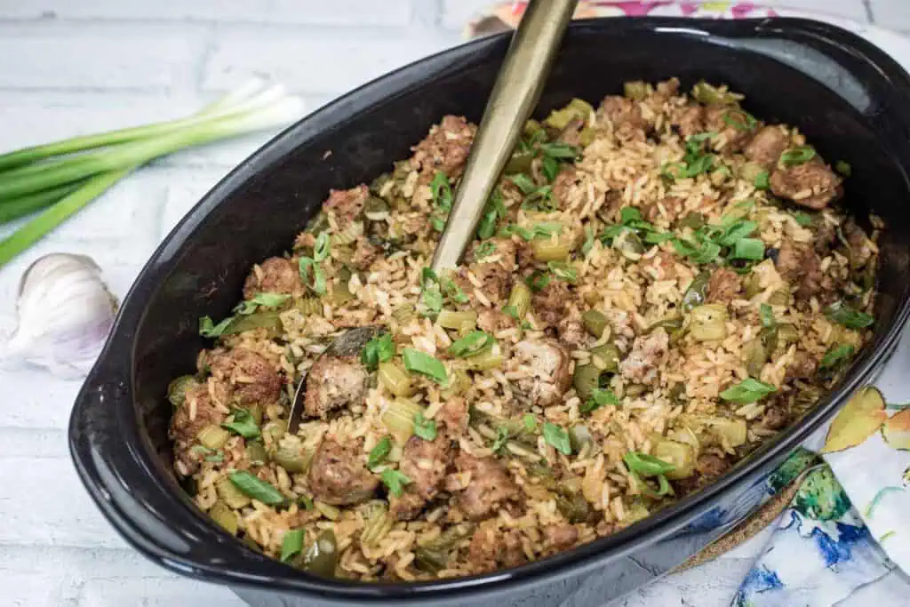 A black casserole dish filled with rice, sausage, and chopped vegetables, garnished with green onions. A serving spoon is placed inside the dish. Scallions and garlic sit nearby.