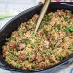 A black casserole dish filled with rice, sausage, and chopped vegetables, garnished with green onions. A serving spoon is placed inside the dish. Scallions and garlic sit nearby.