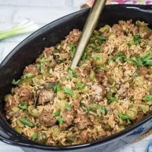 A black casserole dish filled with rice, sausage, and chopped vegetables, garnished with green onions. A serving spoon is placed inside the dish. Scallions and garlic sit nearby.