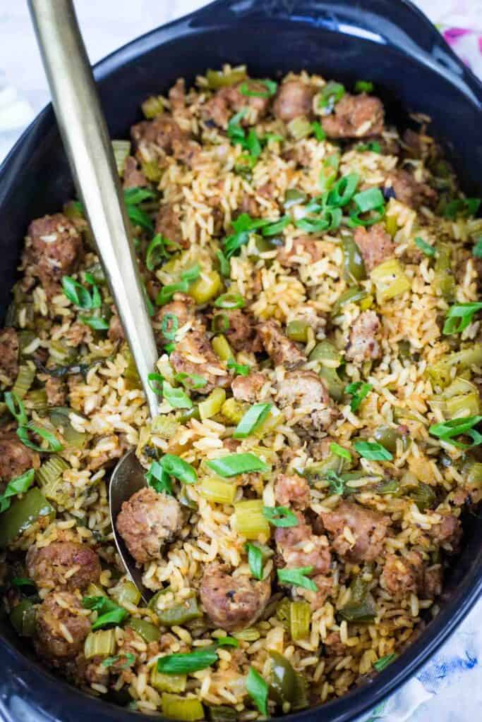 A dish of rice mixed with sausage, celery, green bell peppers, and chopped green onions, served in a black casserole dish with a spoon.