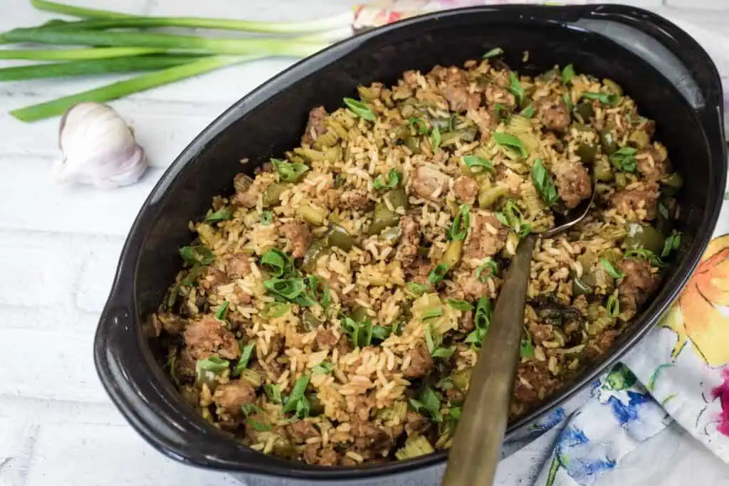 A black oval dish filled with rice, sausage, and chopped green onions sits on a white surface with a spoon, scallions, and garlic nearby.