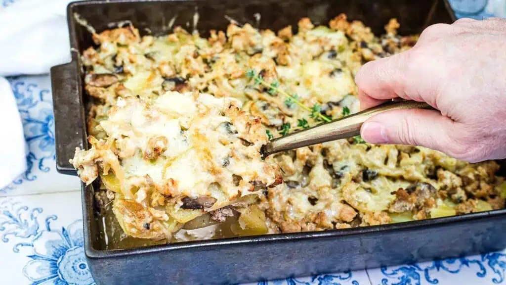 A hand serves a portion of cheesy casserole with visible layers of meat, potatoes, and mushrooms from a rectangular baking dish.