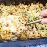 A hand lifts a serving of French Country Ground Pork and Potato Casserole from a rectangular metal pan on a patterned surface.