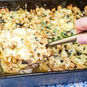 A hand lifts a serving of baked casserole with ground meat, cheese, and vegetables from a rectangular metal pan on a patterned surface.