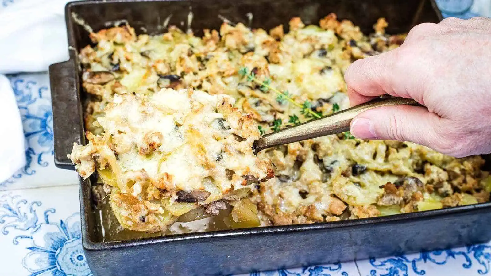 A hand lifts a serving of French Country Ground Pork and Potato Casserole from a rectangular metal pan on a patterned surface.