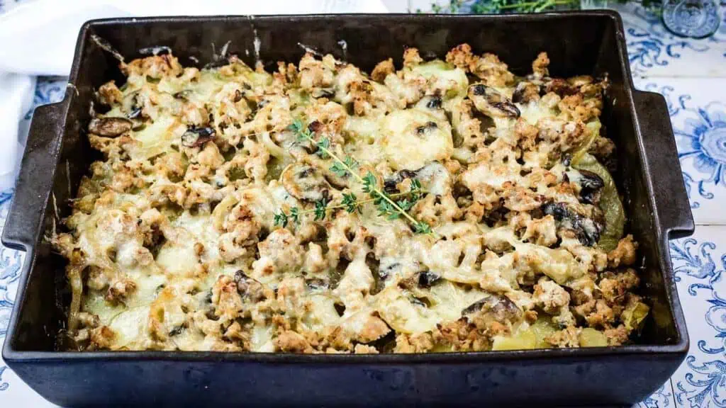 A baked casserole in a dark rectangular dish, topped with browned ground meat, mushrooms, melted cheese, and garnished with fresh thyme.