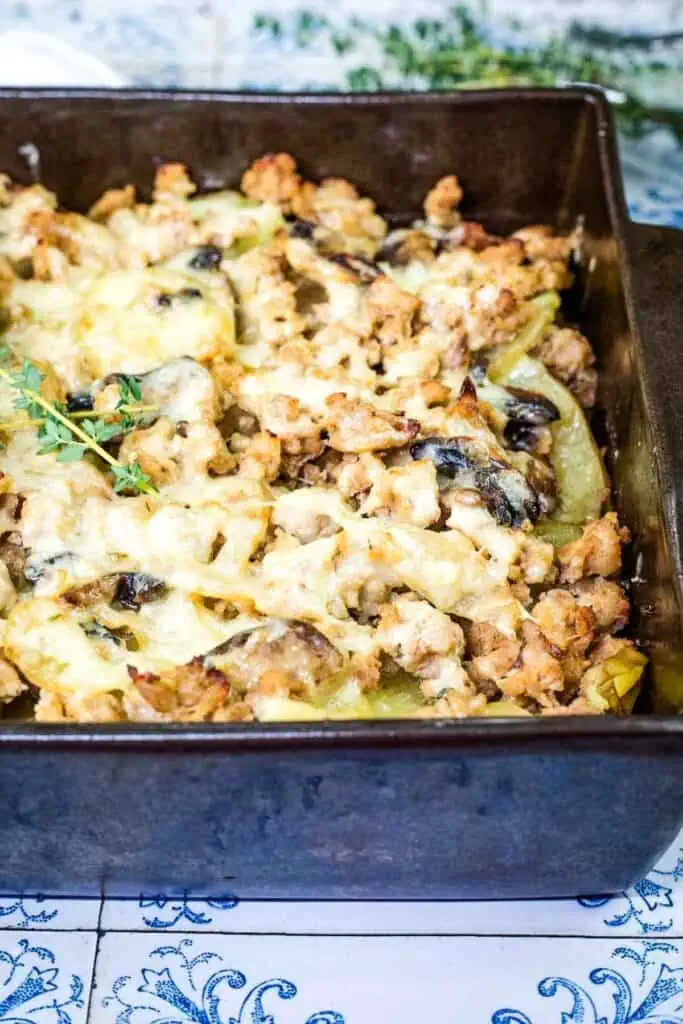 A baked casserole dish filled with layers of sliced vegetables, ground meat, mushrooms, and melted cheese, topped with herbs.
