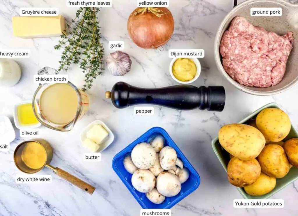 Ingredients for a recipe displayed on a marble surface include ground pork, Yukon Gold potatoes, mushrooms, onion, garlic, cheese, herbs, broth, butter, oil, wine, salt, mustard, and pepper.