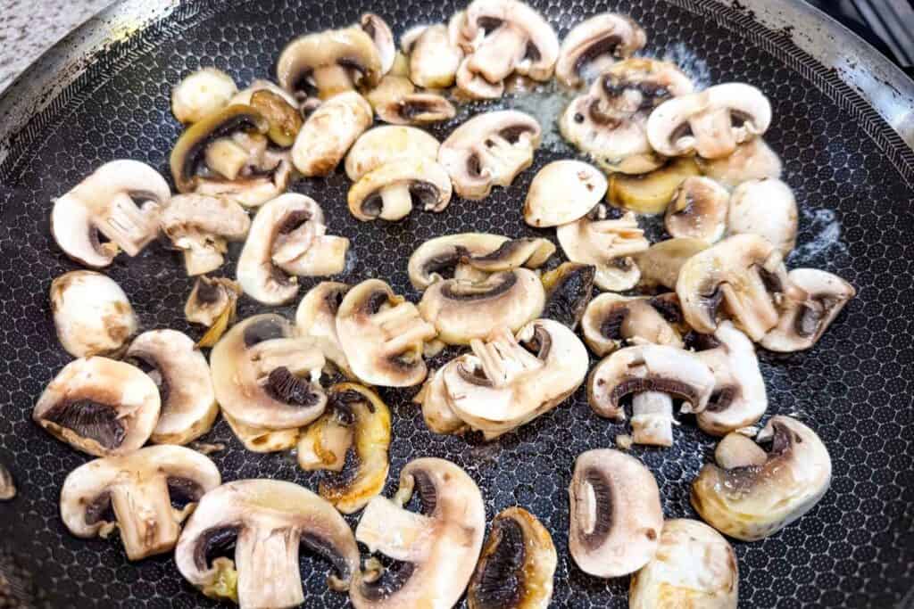 Sliced mushrooms cooking in a frying pan with some browning visible on the edges.
