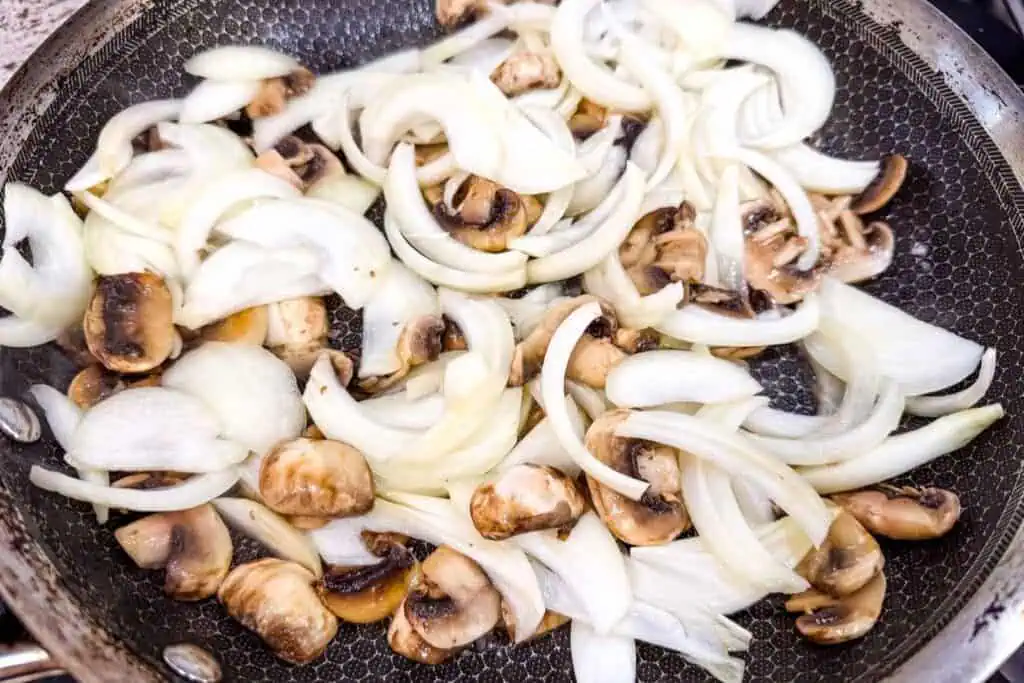 Sliced onions and mushrooms are cooking in a frying pan.