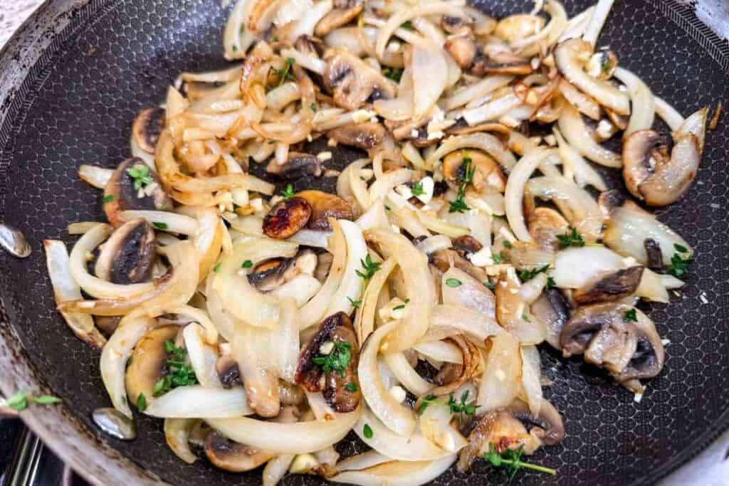Sliced onions and mushrooms sautéing in a black frying pan, garnished with chopped herbs and small pieces of garlic.