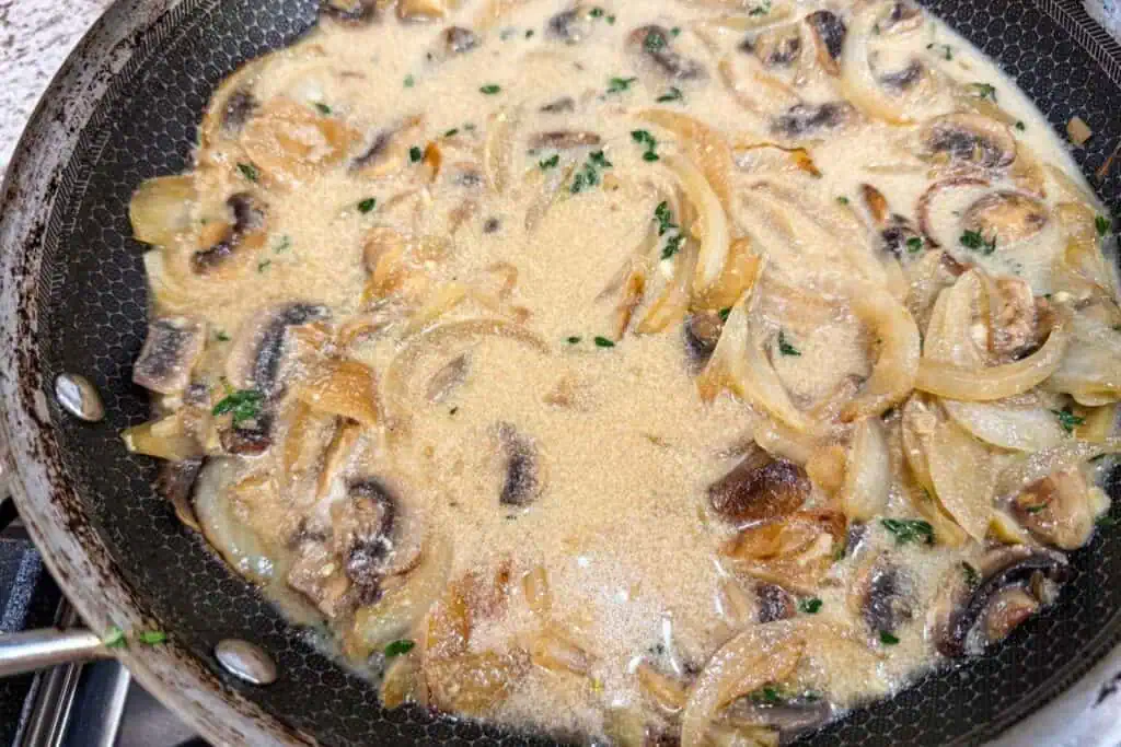 Sautéed onions and mushrooms simmer in a creamy sauce inside a skillet on a stovetop.