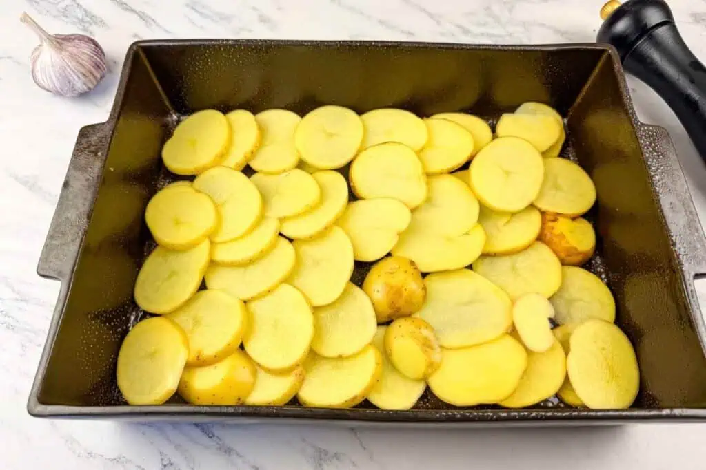 A baking dish filled with a single layer of sliced raw potatoes, placed on a marble countertop next to a garlic bulb and a pepper grinder.