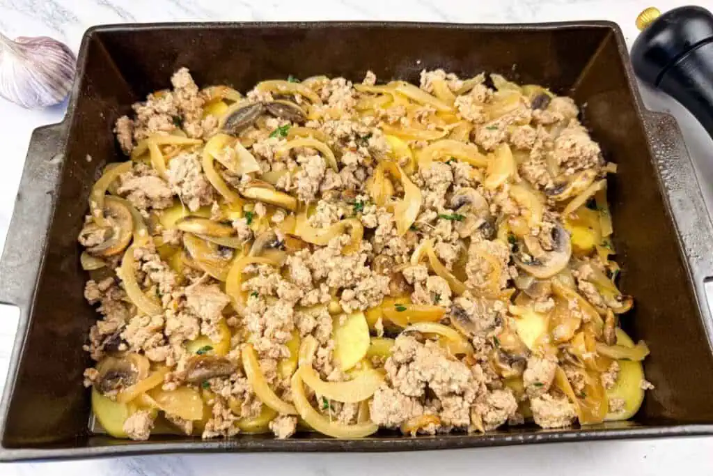 Rectangular baking dish filled with a mixture of cooked ground meat, sliced onions, mushrooms, and herbs on a marble countertop.