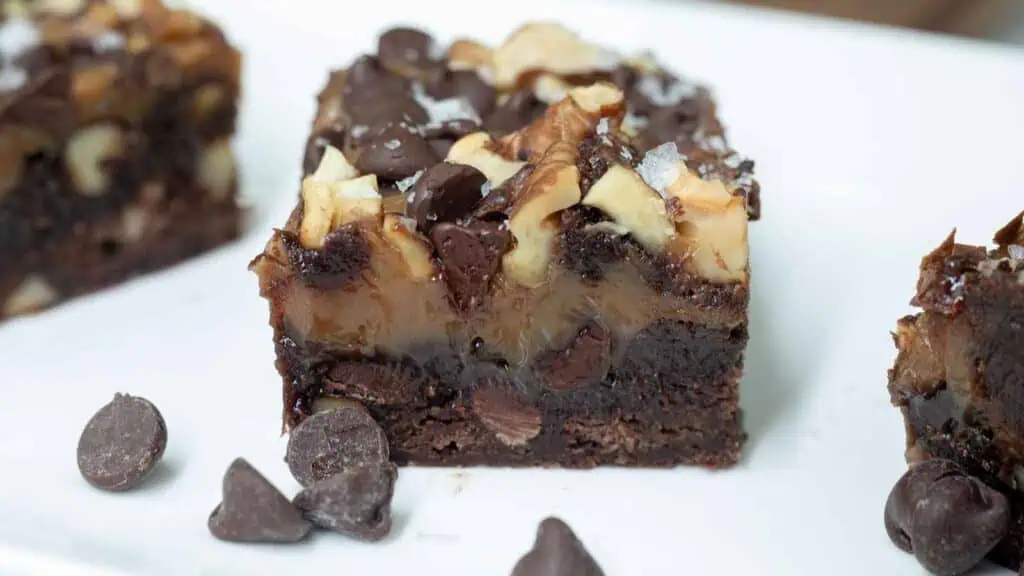 A close-up of a layered chocolate brownie topped with nuts, chocolate chips, and a drizzle of chocolate, on a white plate with extra chocolate chips nearby.