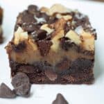 A close-up of a layered chocolate brownie topped with nuts, chocolate chips, and a drizzle of chocolate, on a white plate with extra chocolate chips nearby.