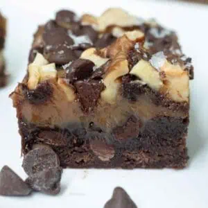 A close-up of a layered chocolate brownie topped with nuts, chocolate chips, and a drizzle of chocolate, on a white plate with extra chocolate chips nearby.