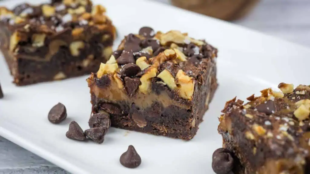 Two chocolate brownies topped with walnuts and chocolate chips on a white plate, with a few chocolate chips scattered around.