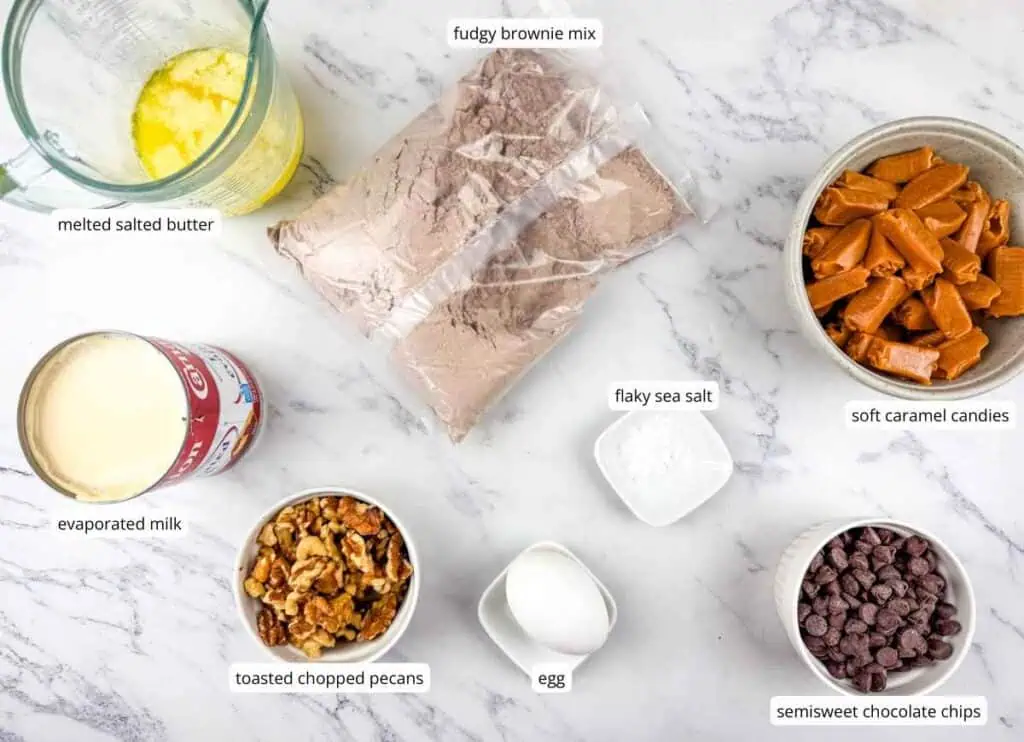Ingredients for a brownie recipe laid out on a white counter: melted butter, brownie mix, caramel candies, evaporated milk, salt, pecans, an egg, and chocolate chips.