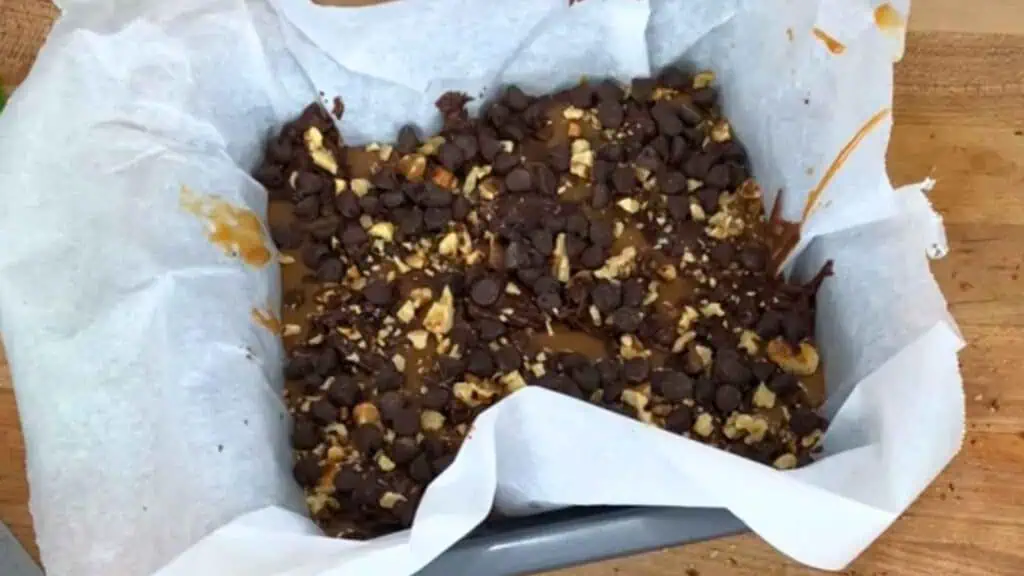 A baking pan lined with parchment paper holds a layered dessert topped with chocolate chips, chopped nuts, and caramel.