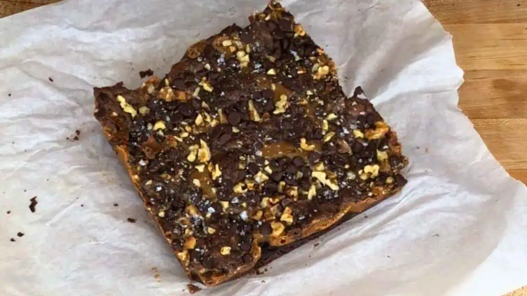 A square chocolate dessert bar with nuts and chocolate chips rests on white parchment paper atop a wooden surface.