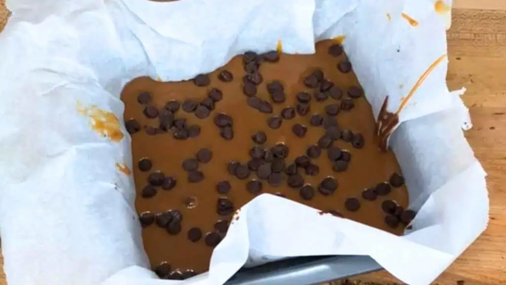 A baking pan lined with parchment paper holds caramel fudge topped with chocolate chips, set on a wooden surface.