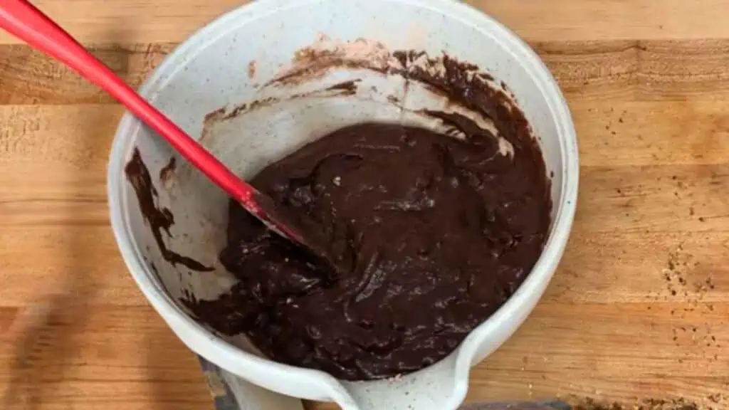 A white mixing bowl with thick chocolate batter and a red spatula sits on a wooden surface.