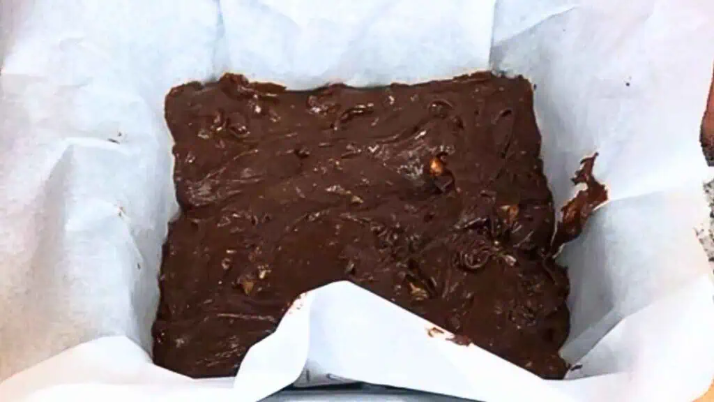 A baking pan lined with parchment paper is filled with unbaked chocolate fudge mixture, spread unevenly and containing visible chunks.