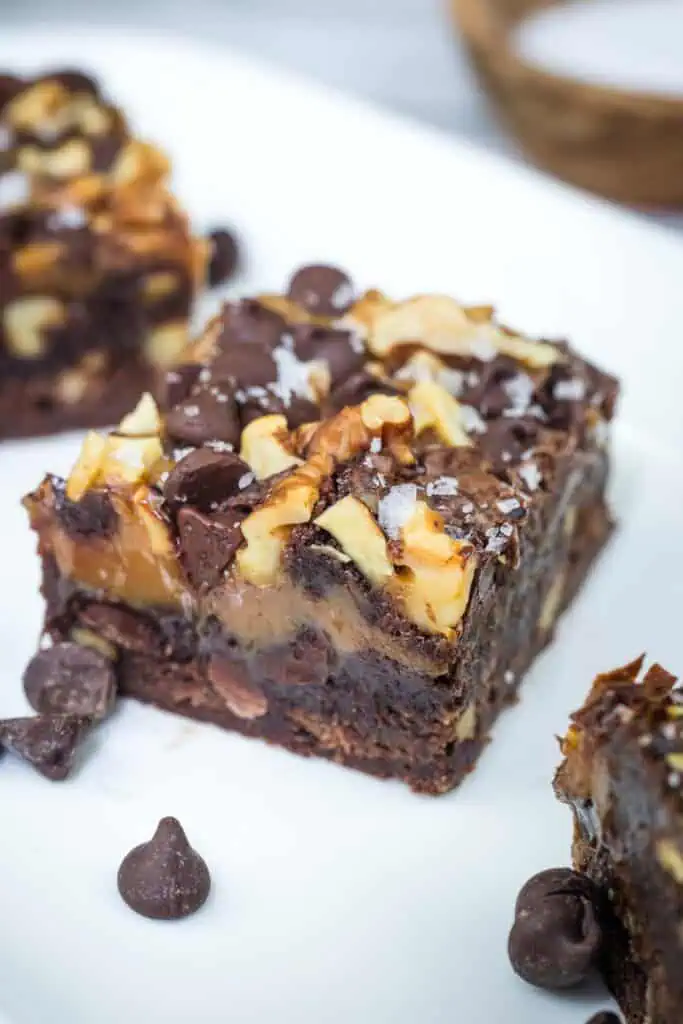 Close-up of a chocolate brownie topped with walnuts, chocolate chips, and a sprinkle of salt, placed on a white plate with a few chocolate chips scattered nearby.