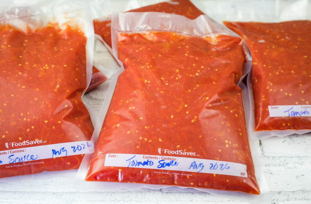 Three vacuum-sealed bags filled with tomato sauce are labeled with the contents and the date “Aug 2026” in blue marker.